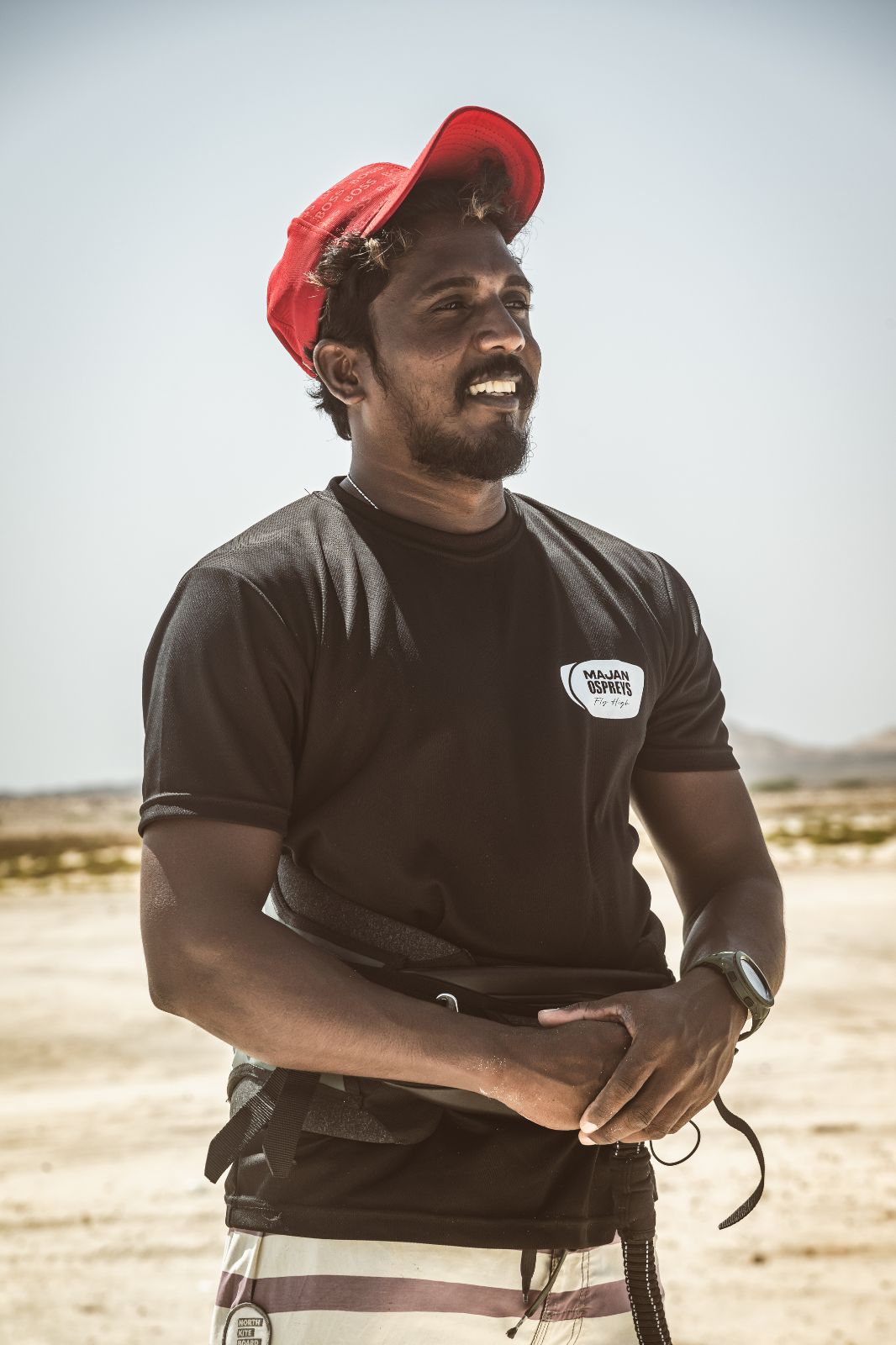 Nalin, a kite instructor, standing on a beach wearing a black polo shirt and red cap, smiling with a kiteboarding backdrop.